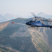 A blue helicopter flies in front of mountain with a long, white boom arm sticking out the front of the aircraft with red tip