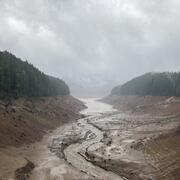 Cloudy rainy day. Pine trees top brown steep muddy banks of empty reservoir with stream running through muddy channel