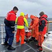 Four people on ship in ocean, huddled around a piece of equipment near railing