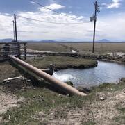 water gushes from a groundwater irrigation well to fill a small channel in a grassy field. Mountains and partly cloudy sky