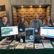 Group of USGS scientists smiling for photo behind booth with handouts and two computer monitors displaying maps