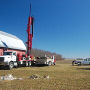 Several vehicles and a large underground drilling rig with a few people in a field beside a building. 