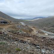 Old buildings, rusted barrel, rocks, and scattered wood from previous buildings in mountain valley. Helicopter near stream.