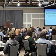 A presenter stands at a podium next to a large screen and speaks to attendees at a conference sitting at round tables