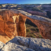red rock arches over a canyon, with shrubs covering nearby rocks and blue sky above