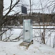 A gray metal streamgage housing with an attached solar panel sits on the shore of a snow-covered frozen river.