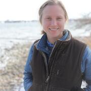A woman with blonde hair in a long sleeved shirt and vest stands for her picture. 