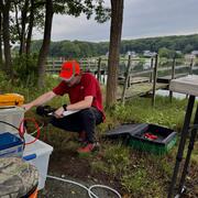 A man wearing a red shirt and orange hat crouches down in front of a peristaltic pump in front of a dock.