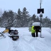 A Hydrologic Technician works on equipment inside a monitoring location housing in knee-deep snow next to a snowmobile