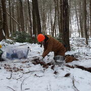 A person with a brown outfit and orange hat takes a soil sample in the woods with snow on the ground.