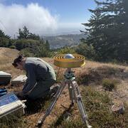 USGS scientist kneels beside gold ring GNSS antenna on a hillside above San Andreas Lake with fog along the distant coast.