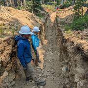 Two people standing in a shallow trench a few feet wide and deep in a forested landscape. They are examining the trench wall