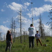 three people stand in a green field, dead trees in background, setting up a tall pole