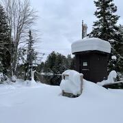 A brown streamgage housing with two feet of fresh snow piled on the roof with a cableway in the background