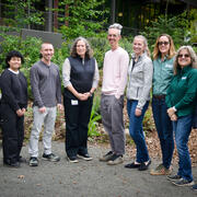 9 people stand in a line with the tallest people in the middle. They are professionally dressed outside amongst greenery