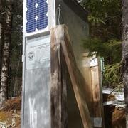 Gray metal USGS streamgage housing with solar panel and inscription mounted on a wooden support in a forested area.
