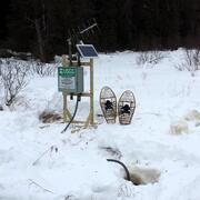 A pair of snowshoes rests in the snow next to a small streamgage mounted on a wooden frame, with antenna and solar panel.