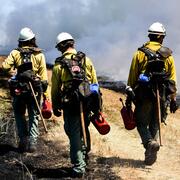 three firefighters walk along a grass trail with materials for implementing prescribed burn, smoke in the background