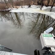 A streamgage housing is mounted on the side of a bridge over a creek. Ice covers the shoreline.