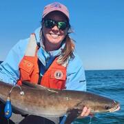 A peson in a hat and sunglasses and orange life jacket leans on the side of a boat holding a cobia