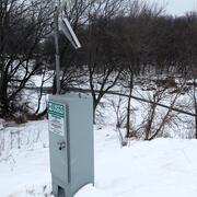 USGS streamgage with solar panel beside a snow-covered riverbank and leafless trees in winter