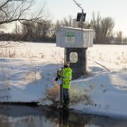 A hydrologic technician wearing a bright yellow coat stands in front of a streamgage housing and takes a streamflow reading