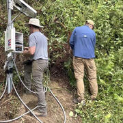 two people working around a landslide monitoring site with thick bushes nearby