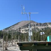 A tall white metal tower with scientific instruments stands on a rooftop of a small building in a mountainous area with scattered rocks and sparse vegetation. In the background, Mammoth Mountain rises as a forested peak under a clear blue sky. The monitoring equipment includes sensors mounted at the top of a 6-meter-high mast for measuring volcanic CO₂ emissions.