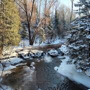 Shallow waterfalls in the middle of a river, surrounded by snow covered trees and foliage on shoreline