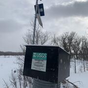 USGS streamgage housing with solar panel and sensors mounted on a pole, against a snow-covered rural background
