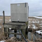 Streamgage housing on wooden platform next to a stream in field in winter