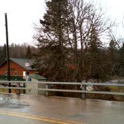 USGS streamgage housing behind bridge guardrails, with a wet road surface on a rural bridge in the foreground.