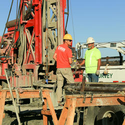 Image shows USGS scientists standing beside a drill rig in protective gear.