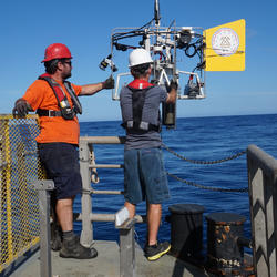USGS scientists stand on the edge of a ship, preparing equipment to go into the ocean