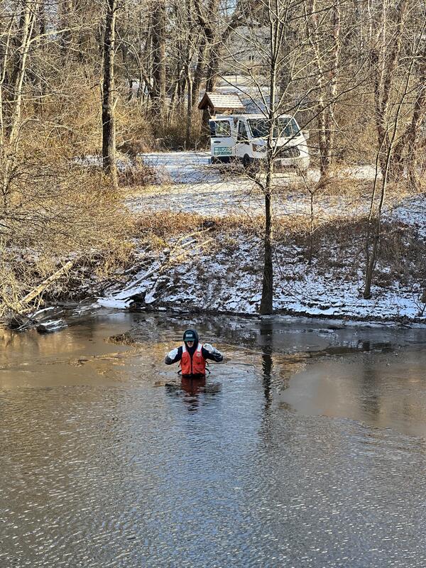 Man in cold weather flotation gear wades into deep river with a snowy bank behind him