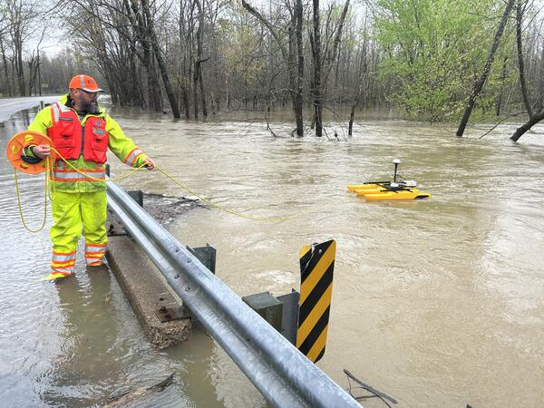 A water expert is wading in ankle-deep floodwaters and holds a rope attached to special equipment that measures water flow. 