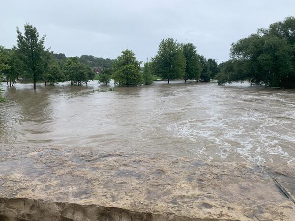 Brown floodwaters swirling in foreground with green trees in background