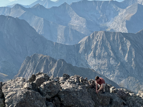 A person is crouched on the ground atop a rocky mountain ridge with numerous ridges seen off in the distance. 
