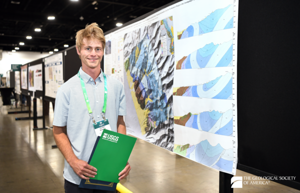 A person stands to the left side of a poster with a colorful geologic map and several cross sections. A long line of posters is seen in the background. 