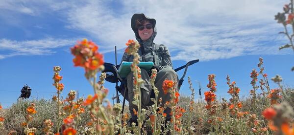 A person sitting in a chair with a clipboard smiles, there are orange globemallow flowers in the foreground and a cloudy blue sky in the background