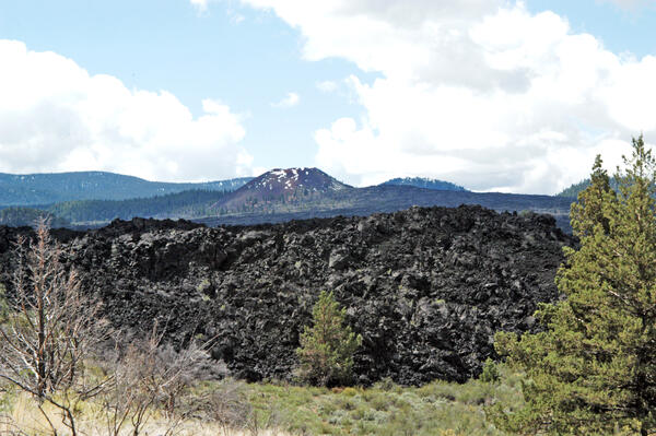 A rubbly black flow of basalt lava, erupted at around 1250°C (2280°F) from the Medicine Lake Volcano. In the distance, the slopes of a scoria cone volcano are covered with similar lava flows and dark pine trees.