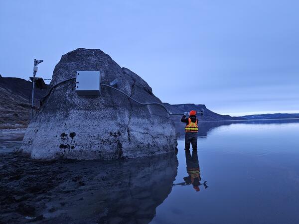 technician in safety gear stands near a large rock amidst the shallow lake waters reflecting a steel-blue early morning sky
