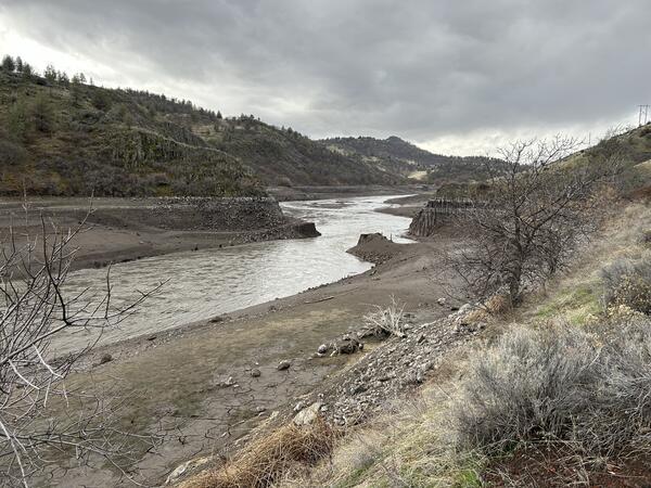 Brown river water flows by muddy banks with course grained sediment. Columns of basalt in right. Cloudy skies.