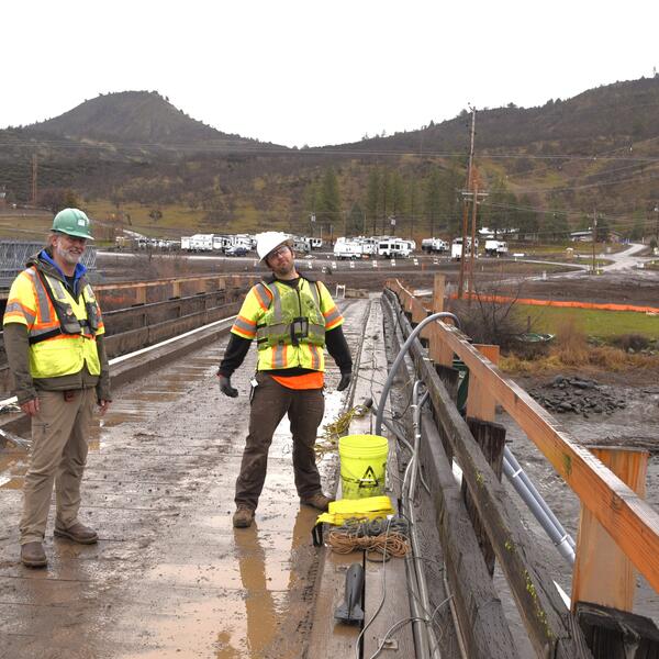 Two scientists in bright colored safety gear and hard hats stand on a bridge above the river.