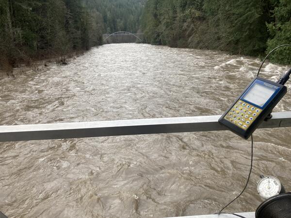 A swollen brown river fills the view. A flow calculator interface is fastened to aluminum rail of cable car.