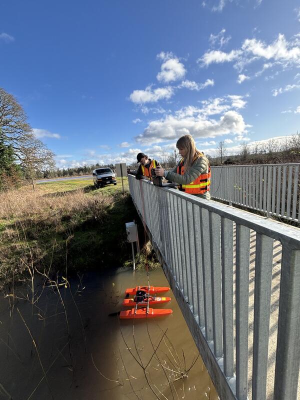two people standing on a foot bridge look over metal railing at measurement equipment in the creek