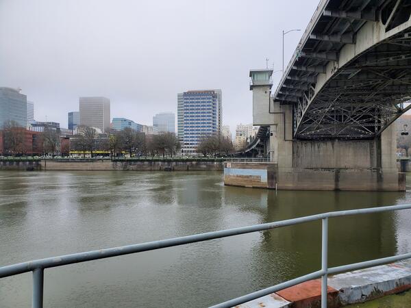 looking at SW Portland waterfront from middle of river. Brown water moves past bridge dolphin. Low clouds shroud skyscrapers