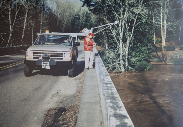 Man in red float coat operates a truck mounted crane to lower equipment into the dark brown river water