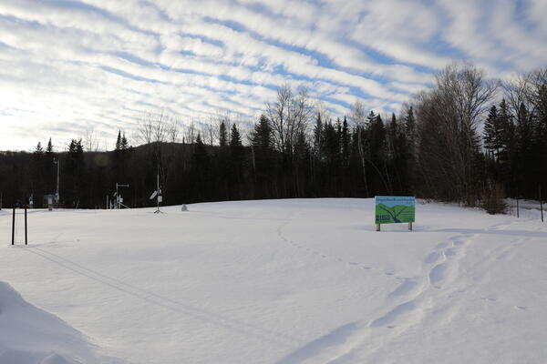 A snowy field with a blue and green sign on it and some meteorological equipment.