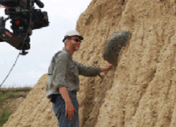 A geologist stands at an outcrop containing a small boulder deposited by a glacier with a video camera in the foreground. 